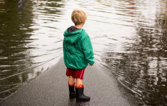 Kleiner Junge mit Regenjacke und Gummistiefeln steht auf einer überschwemmten Straße.
