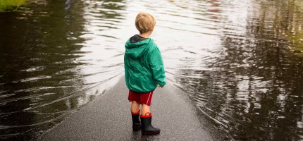 Kleiner Junge mit Regenjacke und Gummistiefeln steht auf einer überschwemmten Straße.