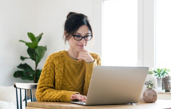 Frau mit gelbem Strickpulli und Brille sitzt vor einem Laptop.