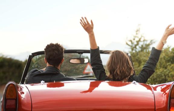 Zwei Personen sitzen in einem roten Cabrio, während sie auf einer Straße fahren. Die Person auf der Beifahrerseite hat die Arme freudig in die Luft gestreckt.