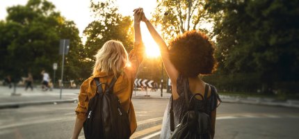 Zwei Frauen halten bei Sonnenuntergang Händchen und recken die Arme in die Luft.