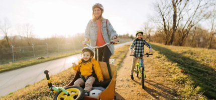 Eine Mutter mit zwei Kindern fährt Lastenfahrrad. Ein Kind fährt mit einem Kinderrad nebenher, das zweite sitzt im Lastenrad.