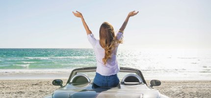 Eine Frau sitzt auf der Rücksitzbank ihres Cabrios am Strand und wirft ihre Arme in die Höhe während Sie aufs Meer schaut.