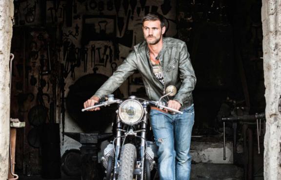 Ein Mann in Lederjacke und Jeans schiebt ein Motorrad aus einer Scheune.
