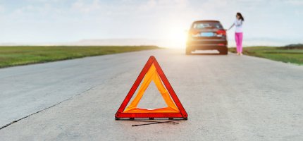 Ein Warndreieck steht auf einer Landstraße hinter einem Auto neben dem eine Frau steht.