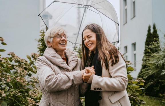 Eine alte und eine junge Frau stehen unter einem Regenschirm und lachen.