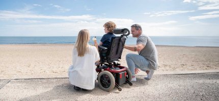 Elternpaar mit einem Kind im Elektrorollstuhl am Strand.