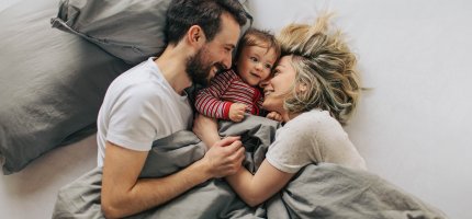 Paar liegt glücklich im Bett mit Kleinkind zwischen sich.