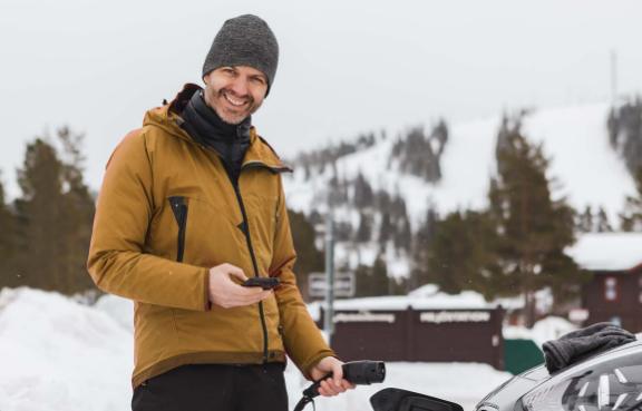 Person lädt ein Elektroauto an einer Ladestation in winterlicher Berglandschaft, hält ein Smartphone; im Hintergrund schneebedeckte Hügel, Bäume und Hütten.