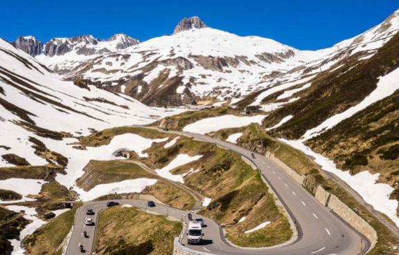 Eine kurvenreiche Straße führt durch eine beeindruckende Berglandschaft mit schneebedeckten Gipfeln. Mehrere Fahrzeuge sind auf der Straße unterwegs.