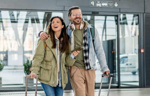Ein Paar mit Rollkoffern und Smartphone steht vor einem modernen Gebäude, bereit für die Reise.