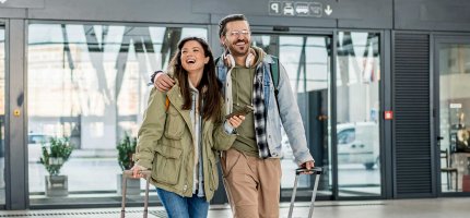 Ein Paar mit Rollkoffern und Smartphone steht vor einem modernen Gebäude, bereit für die Reise.