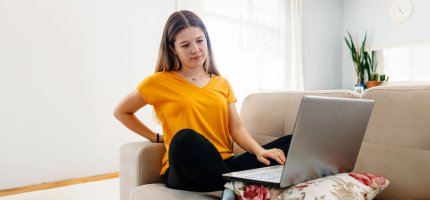 Junge Frau sitzt auf dem Sofa vor dem Laptop und hält sich den Rücken.