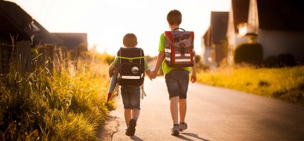 Zwei kleine Jungs mit Schulranzen gehen Hand in Hand durch ein Dorf.