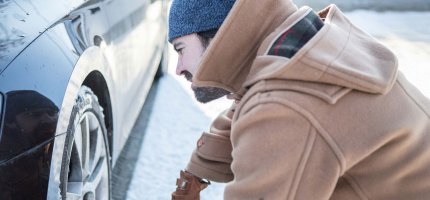 Eine Person trägt warme Kleidung und überprüft den Reifen eines Autos im Schnee.