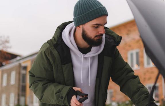 Eine Person steht draußen im Schnee und hält ein Starthilfekabel, während sie am offenen Kofferraum eines Autos arbeitet.