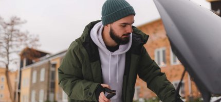 Eine Person steht draußen im Schnee und hält ein Starthilfekabel, während sie am offenen Kofferraum eines Autos arbeitet.