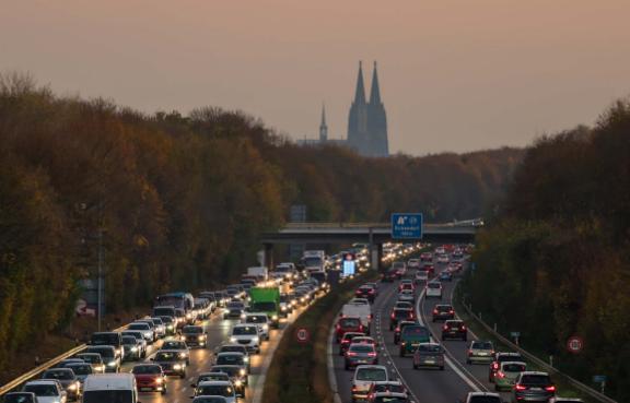 Dichter Verkehr auf einer Autobahn bei Sonnenuntergang, mit einer Stadtlandschaft im Hintergrund, einschließlich einer markanten Kathedrale.