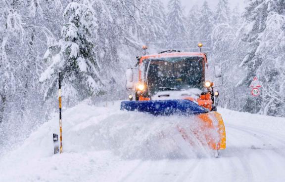 Ein Schneepflug räumt eine verschneite Straße, umgeben von schneebedeckten Bäumen.