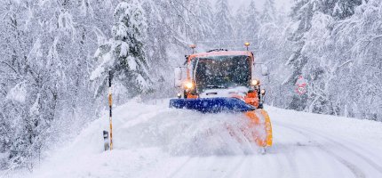 Ein Schneepflug räumt eine verschneite Straße, umgeben von schneebedeckten Bäumen.