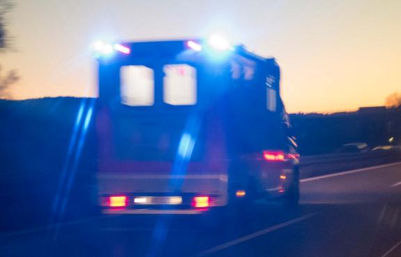 Rettungswagen mit blauem Blaulicht fährt bei Dämmerung über eine Landstraße, andere Fahrzeuge im Hintergrund.