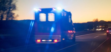 Rettungswagen mit blauem Blaulicht fährt bei Dämmerung über eine Landstraße, andere Fahrzeuge im Hintergrund.
