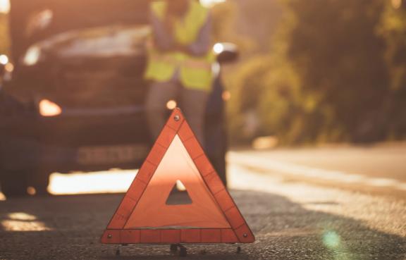 Eine Person lehnt an einem Auto, die Motorhaube ist geöffnet, im Vordergrund steht ein Warndreieck.