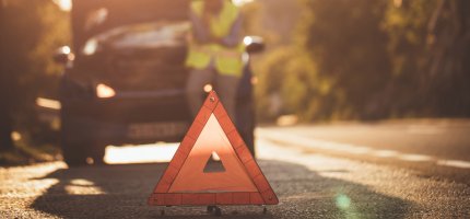 Eine Person lehnt an einem Auto, die Motorhaube ist geöffnet, im Vordergrund steht ein Warndreieck.