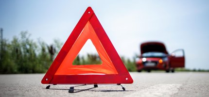 Tragbares Warndreieck auf der Straße soll auf das Pannenfahrzeug im Hintergrund aufmerksam machen.