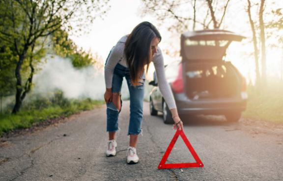 Eine Frau stellt auf einer Straße durch ein Waldstück ein Warndreieck hinter ihrem Auto auf.