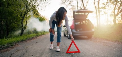 Eine Frau stellt auf einer Straße durch ein Waldstück ein Warndreieck hinter ihrem Auto auf.