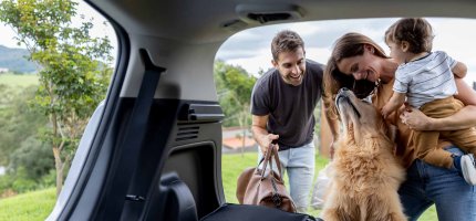 Ein Mann und eine Frau stehen mit einem Kleinkind und einem großen Hund vor einem geöffneten Kofferraum.