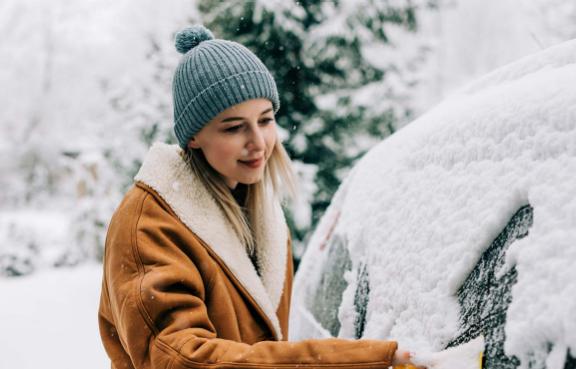 Eine Person entfernt Schnee von der Windschutzscheibe eines Autos in einer verschneiten Umgebung.