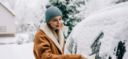 Eine Person entfernt Schnee von der Windschutzscheibe eines Autos in einer verschneiten Umgebung.