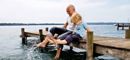 Großvater sitzt mit Enkel auf einem Steg am See und plantscht mit den Füßen im Wasser.