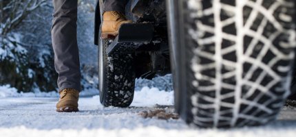 Nahaufnahme von Winterreifen auf einer schneebedeckten Straße, daneben steht eine Person mit Stiefeln.