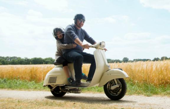 Ein Mann fährt mit einem Motorroller an einem Weizenfeld vorbei. Sein kleiner Sohn umarmt ihn von hinten.