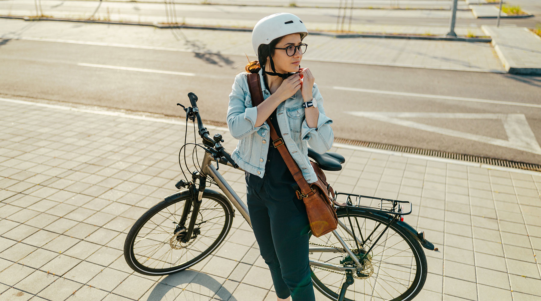 Junge Frau in Jeansjacke und mit Umhängetasche steht vor einem Fahrrad und zieht sich einen Fahrradhelm auf.