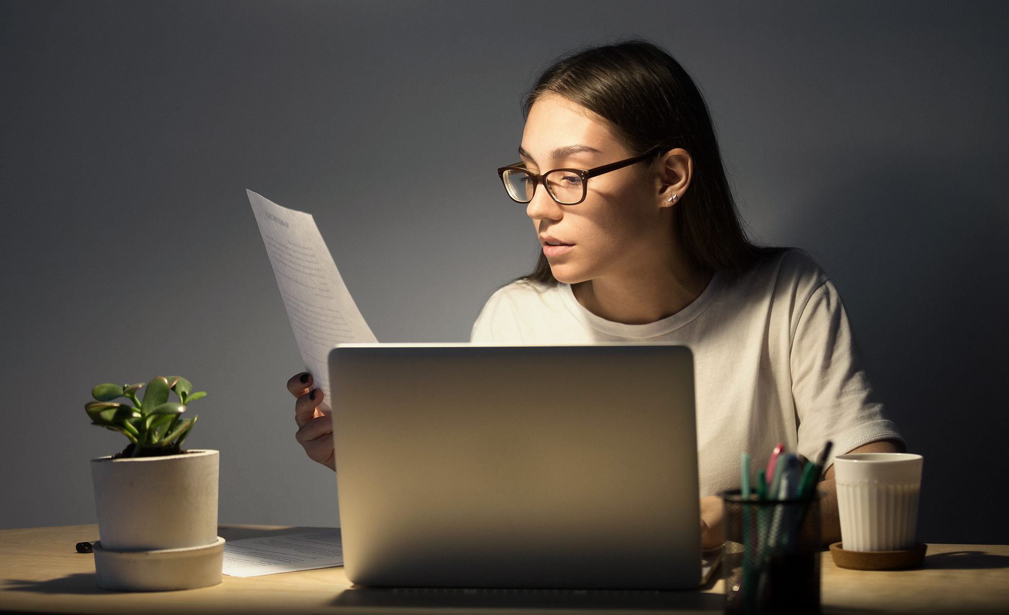 Junge Frau mit Brille liest am Laptop ein Dokument, das sie in der rechten Hand hält.