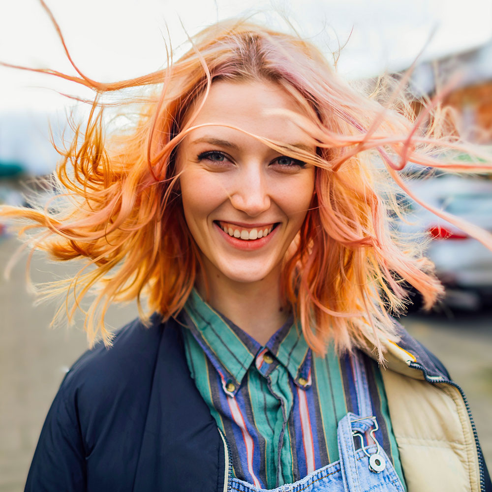 Eine Person steht draußen, ihre Haare wehen im Wind. Sie trägt eine gestreifte Bluse und eine Jacke. Im Hintergrund sind Autos zu sehen.