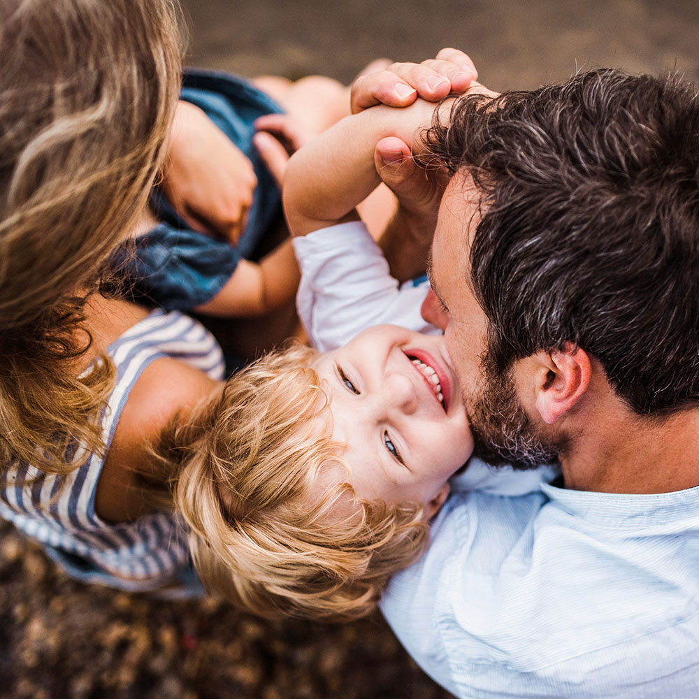 Eine Familie sitzt zusammen, die Eltern halten ein kleines Kind. Sie sind eng beieinander und tragen sommerliche Kleidung. Der Blickwinkel ist von oben.