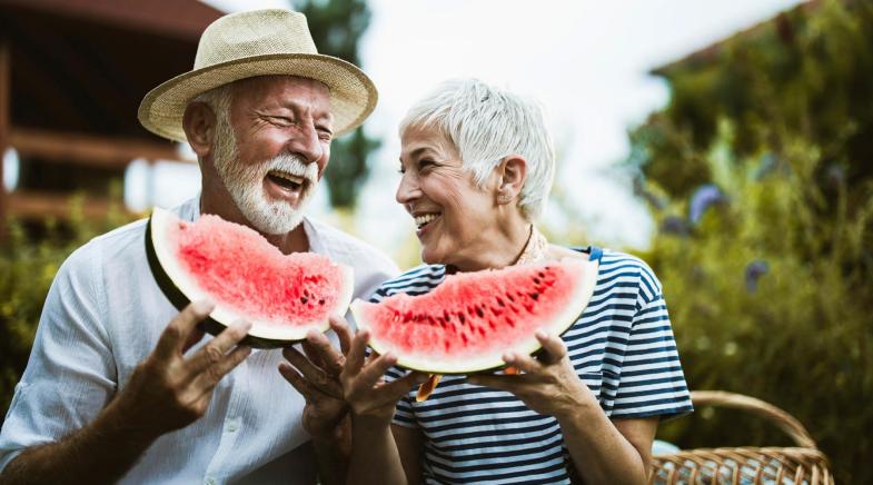 Rentnerpärchen hält zwei Melonenstücke in den Händen, schaut sich an und lacht.