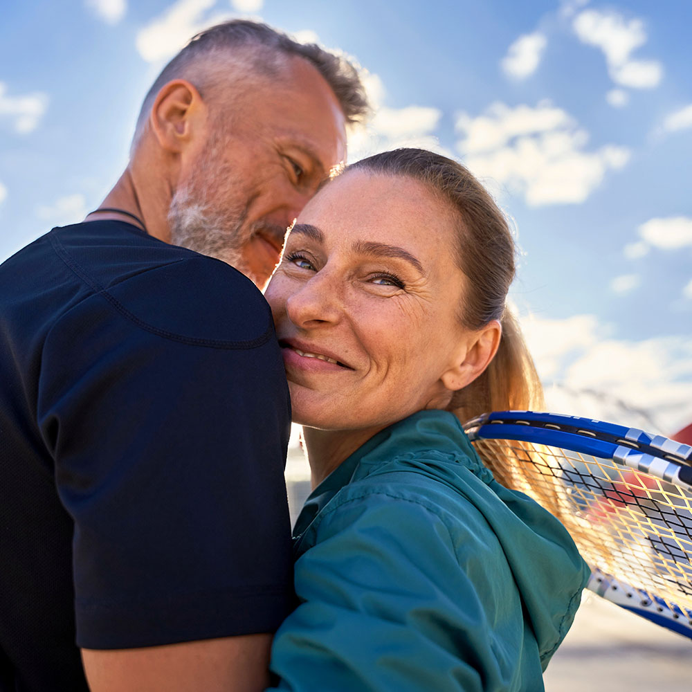 Ein Paar steht draußen, die Frau trägt einen Tennisschläger auf der Schulter. Der Himmel ist klar und sonnig mit einigen Wolken.