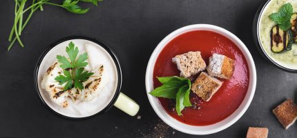 Man schaut von oben auf einen Tisch mit zwei Suppentassen und einer Suppenschüssel. In der linken Suppentasse ist eine Brokkolicremesuppe, in der Suppenschüssel eine Rotebeetecremesuppe und in der rechten Suppentasse eine Zucchinicremesuppe