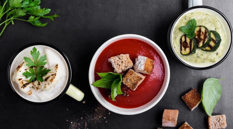 Man schaut von oben auf einen Tisch mit zwei Suppentassen und einer Suppenschüssel. In der linken Suppentasse ist eine Brokkolicremesuppe, in der Suppenschüssel eine Rotebeetecremesuppe und in der rechten Suppentasse eine Zucchinicremesuppe.