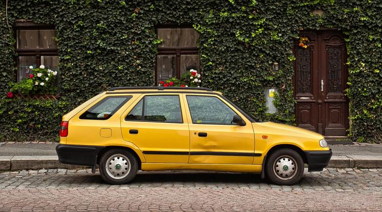 Ein alter Kombi mit Dellen steht am Straßenrand vor einem Haus, dessen Fassade mit Pflanzen bewachsen ist.