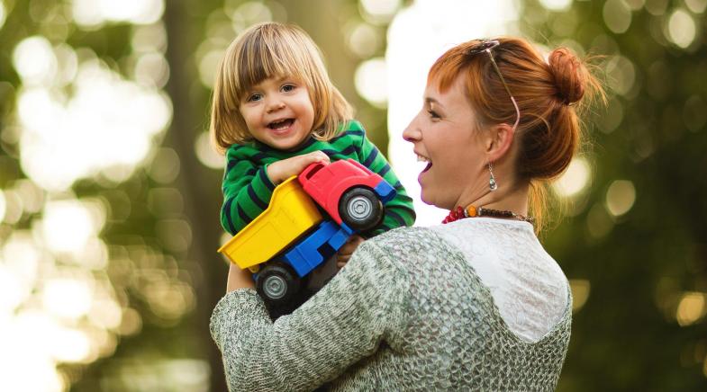 Eine Frau hält lachend ein Kleinkind hoch, das einen bunten Spielzeuglastwagen in der Hand hält.