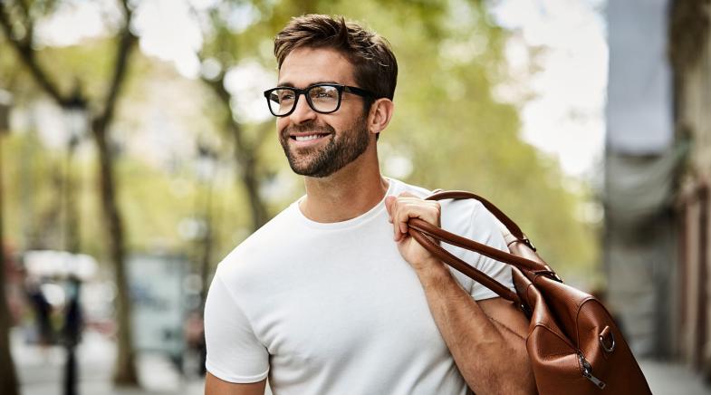 Mann mit Brille läuft mit einer Tasche über der Schulter lächelnd durch die Stadt.