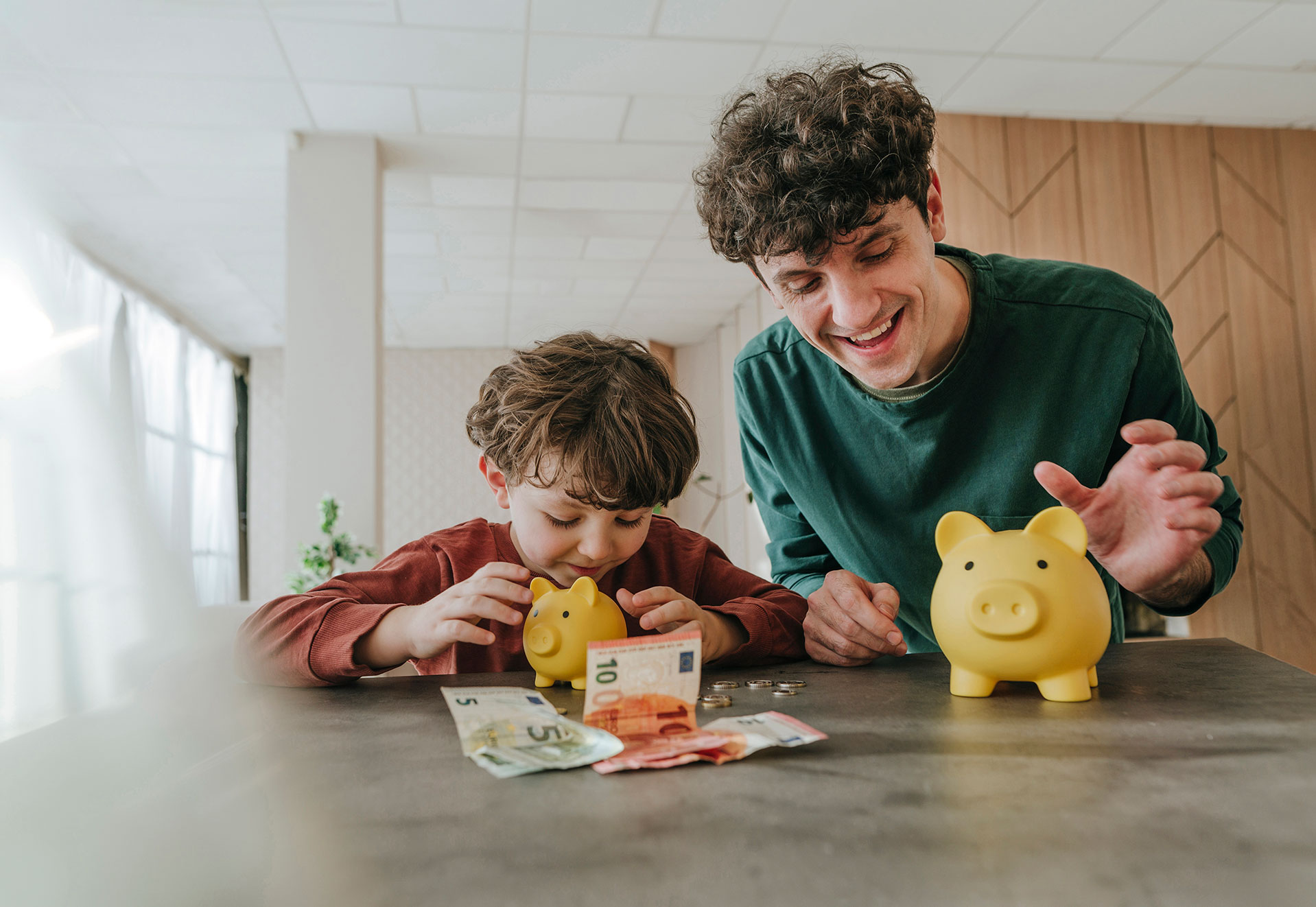 Vater und Sohn sitzen lächelnd am Tisch auf dem Geld und 2 Sparschweine liegen.