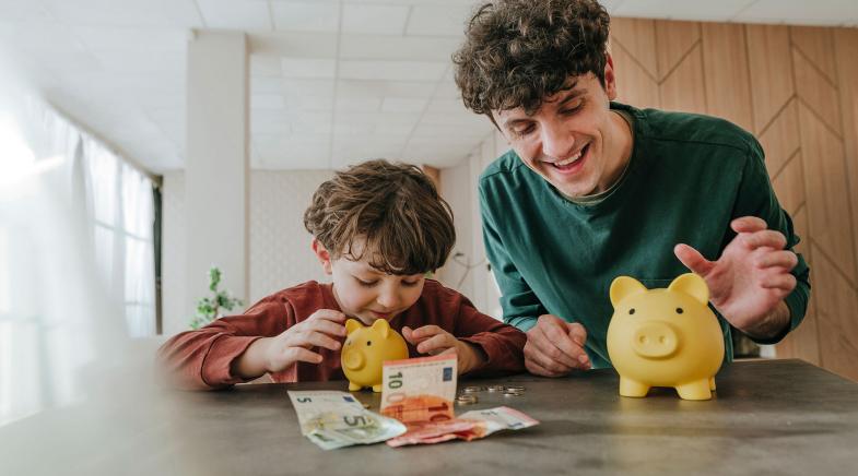 Vater und Sohn sitzen lächelnd am Tisch auf dem Geld und 2 Sparschweine liegen.